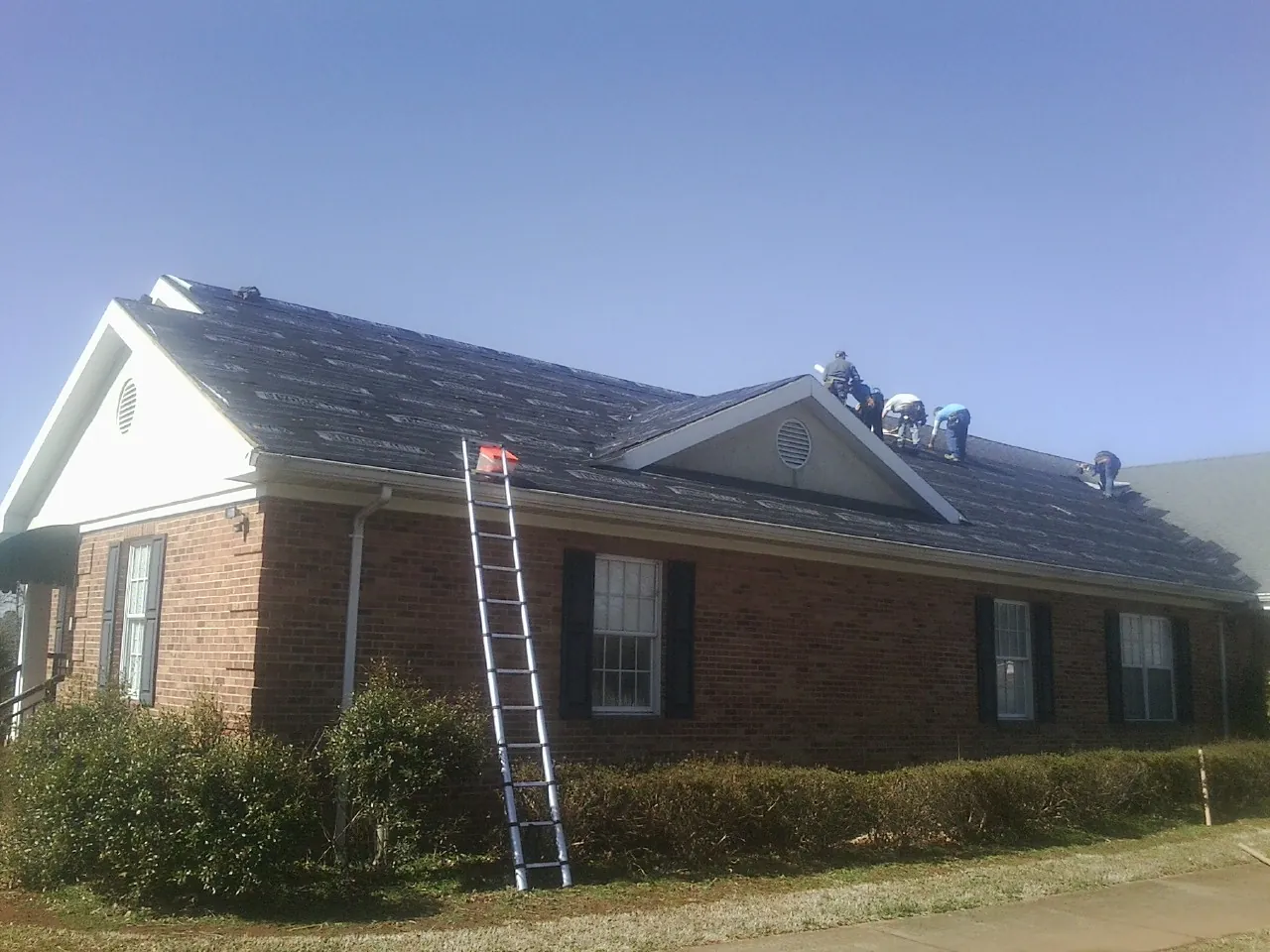 Crew tearing shingles off a brick ranch roof with a ladder set — Emergency Roof Repair work by our crew in Gates