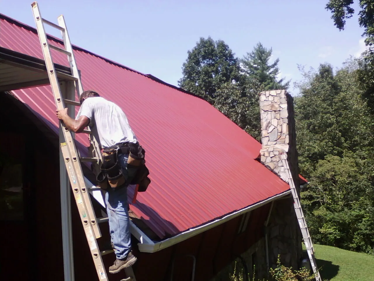 Roofer on a ladder beside a red metal roof and stone chimney — Soffit & Fascia Repair by Roofing Brothers in Gates